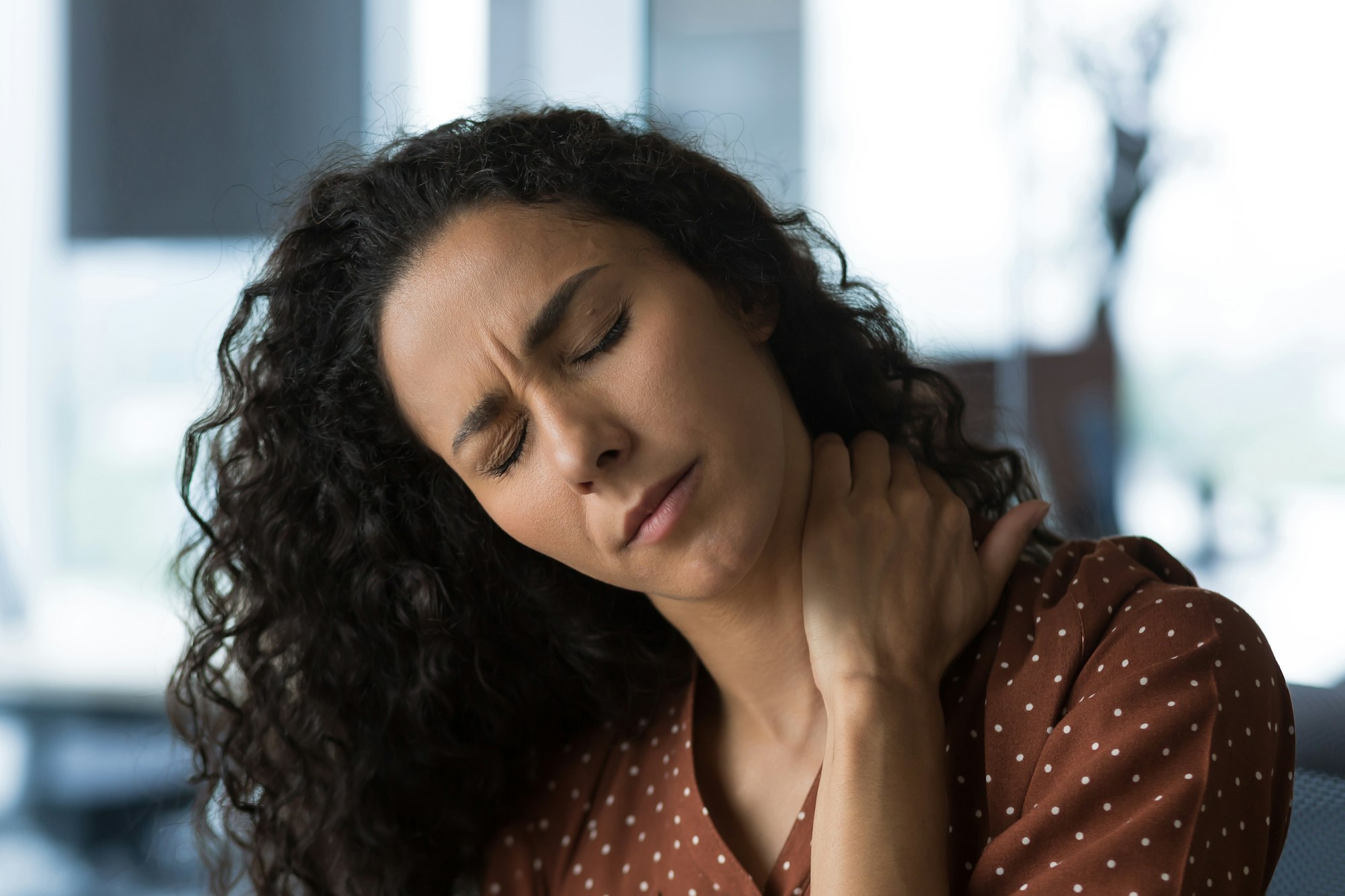 Close-up photo of curly-haired woman at home near the window with severe neck pain